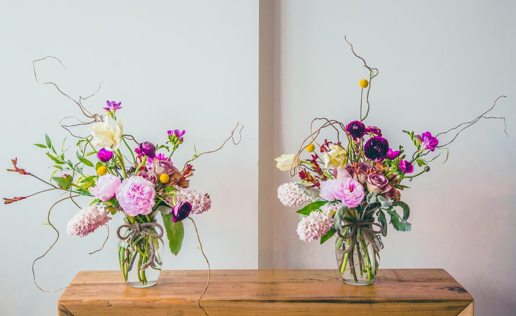 Two loose bouquets of pink, purple and white flowers in glass jars on a rustic wooden bench