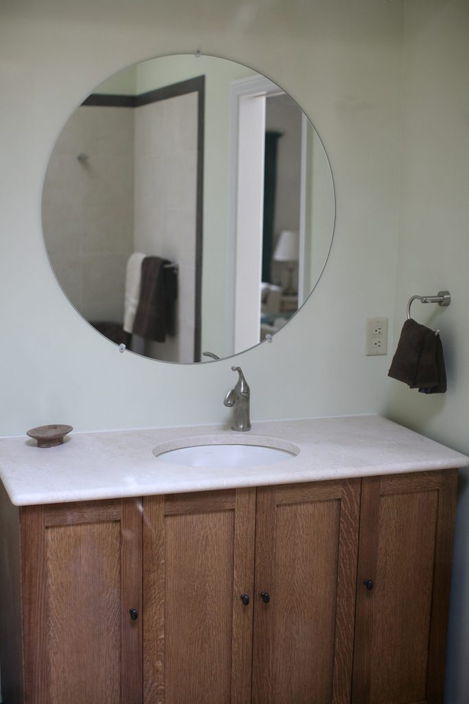 A large round mirror hung above an oak vanity with a white stone top